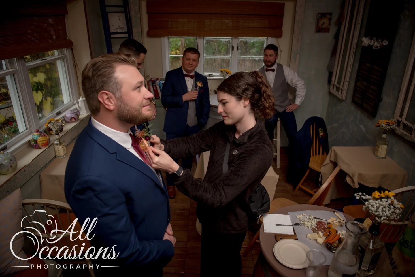Serena carefully pinning a boutonnière to a groom’s lapel while the wedding party gets ready.
