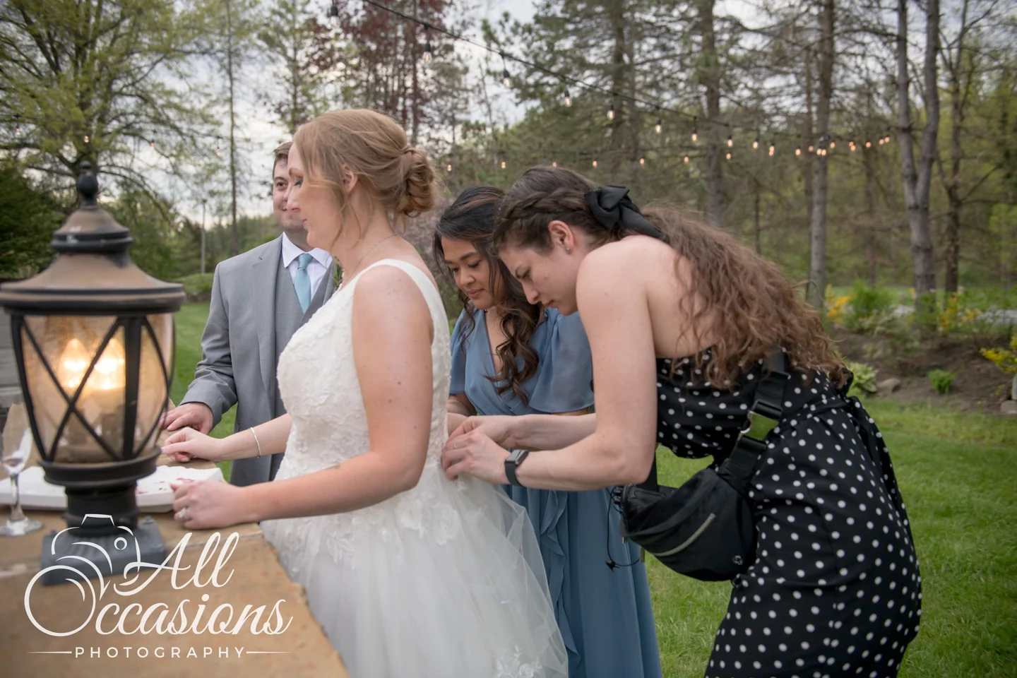 Serena and another member of the wedding party helping with the back of a bride’s dress outdoors.