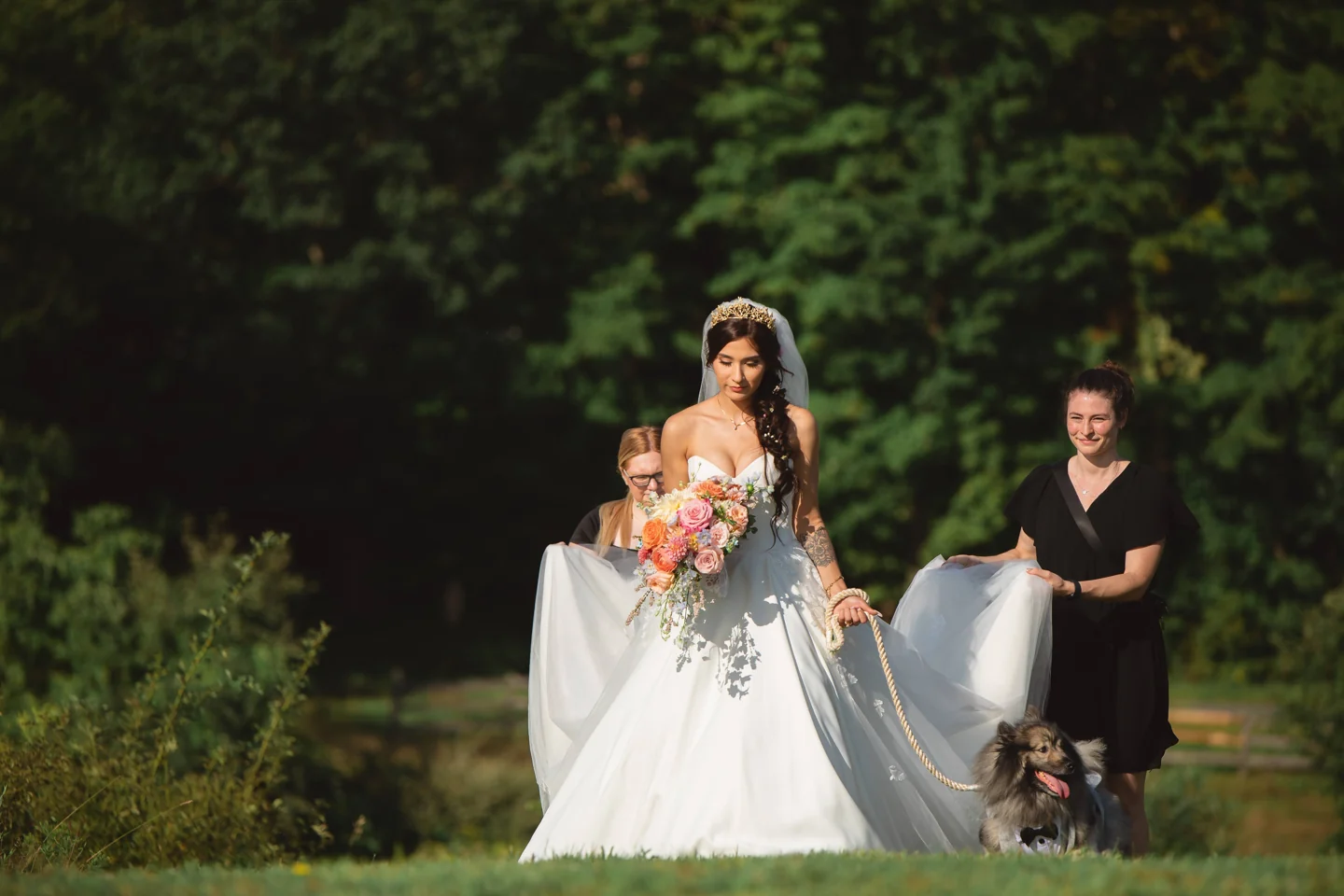 Serena helping carry a bride’s dress outdoors as the bride walks with her dog.